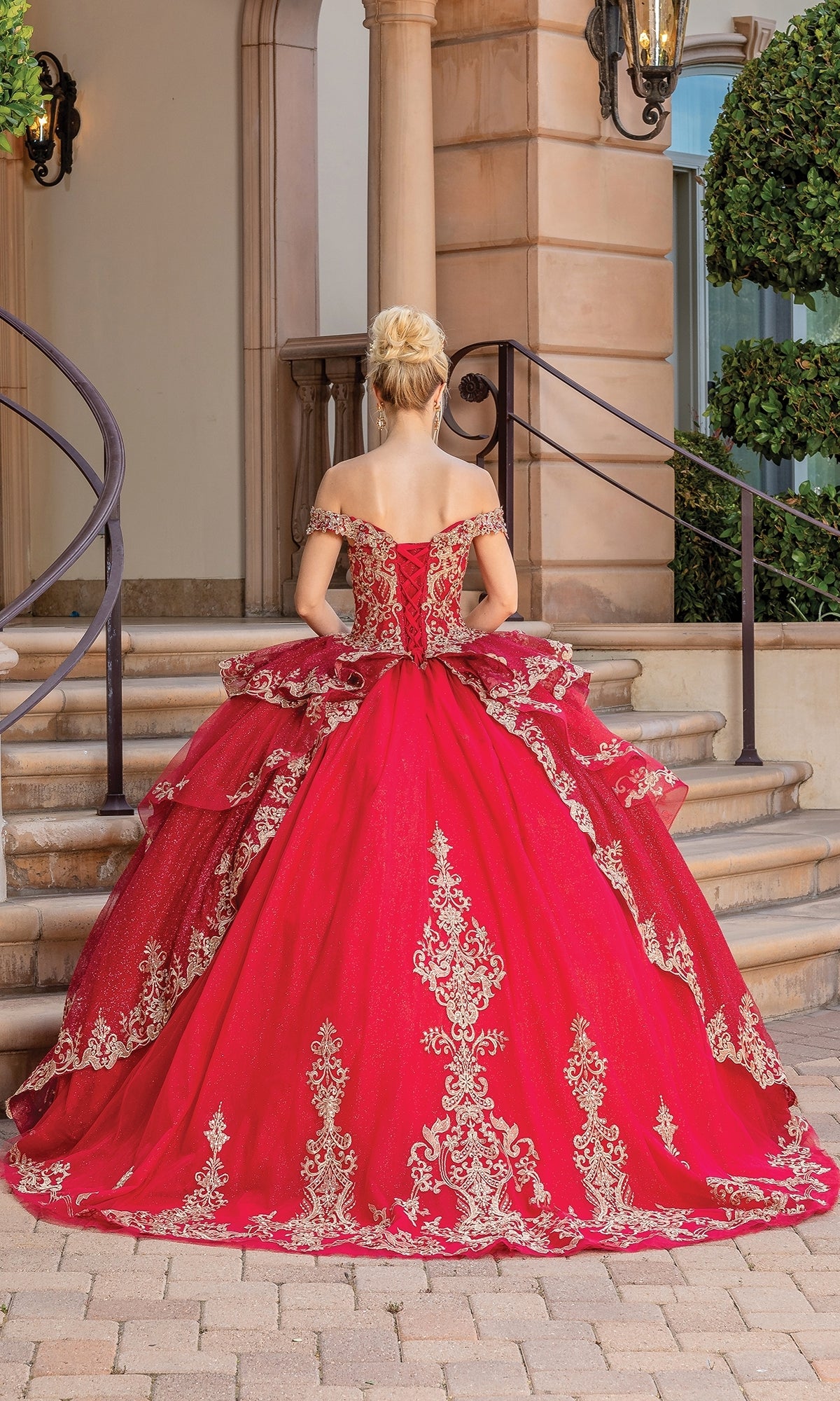 A woman wearing the Quinceanera Dress 1720 By Dancing Queen, an off-the-shoulder gold ball gown with lace embroidery, stands on a cobblestone path before stone steps and ornate railings, facing away from the camera.
